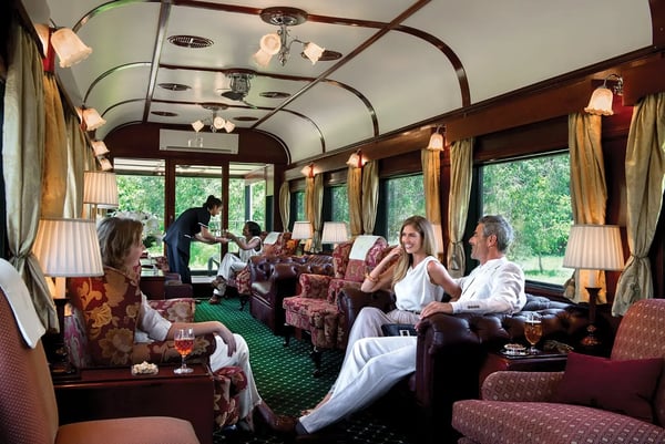 Reisende sitzen in einem eleganten Salonwagen mit Sesseln, Lampen und grossen Fenstern, während Service angeboten wird