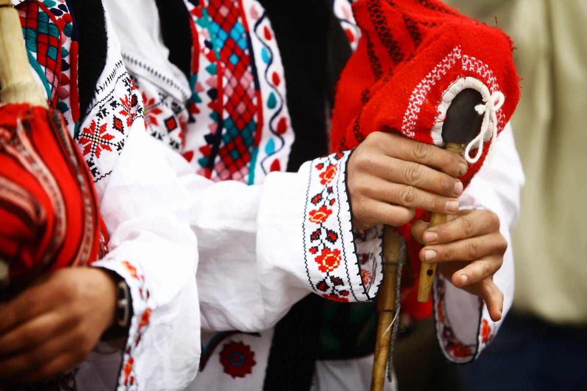 Nahaufnahme von Händen in bestickter Tracht, die eine rote, verzierte Tasche und eine Holzflöte halten