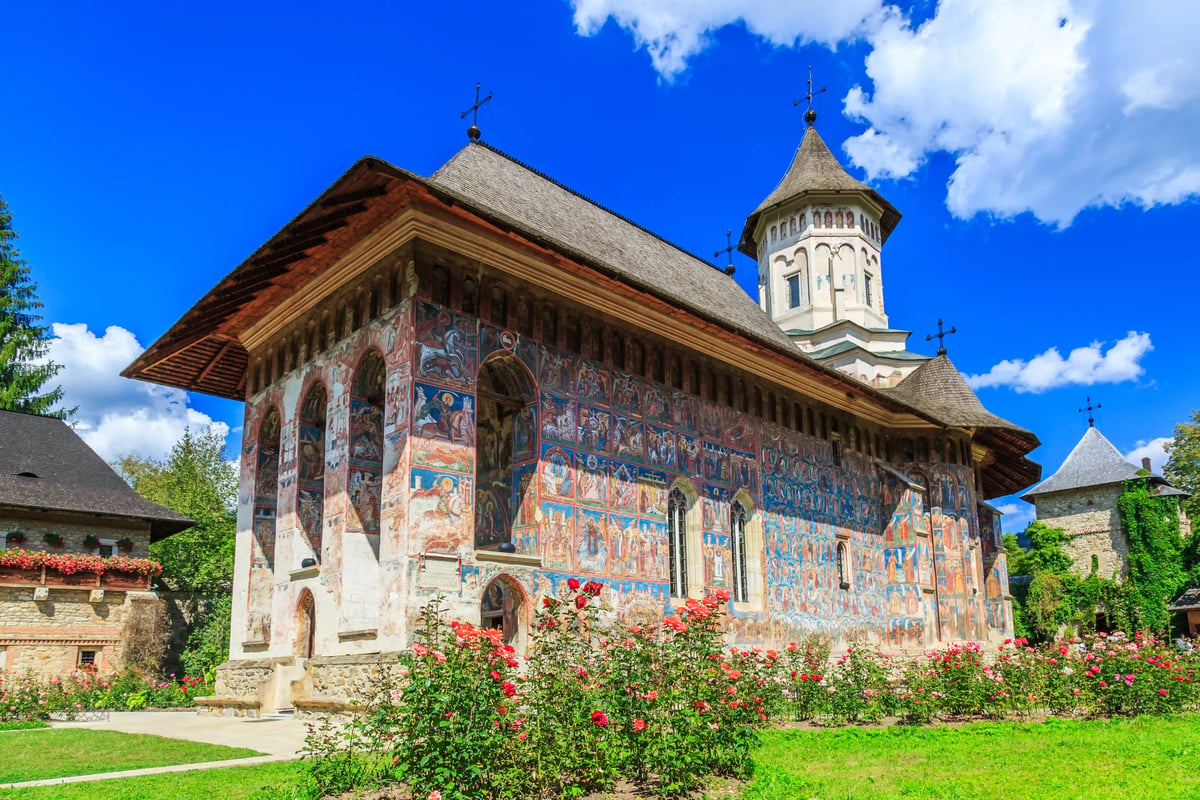 Aussenansicht des UNESCO-Klosters Moldovita mit farbigen Fresken, Kirchturm und Rosenbeeten vor blauem Himmel