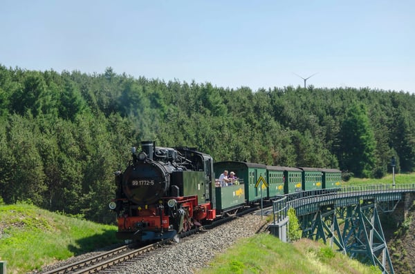 Historische Dampfeisenbahn fährt über eine Stahlbrücke durch grünes Hügelland mit Fahrgästen in offenen Wagen