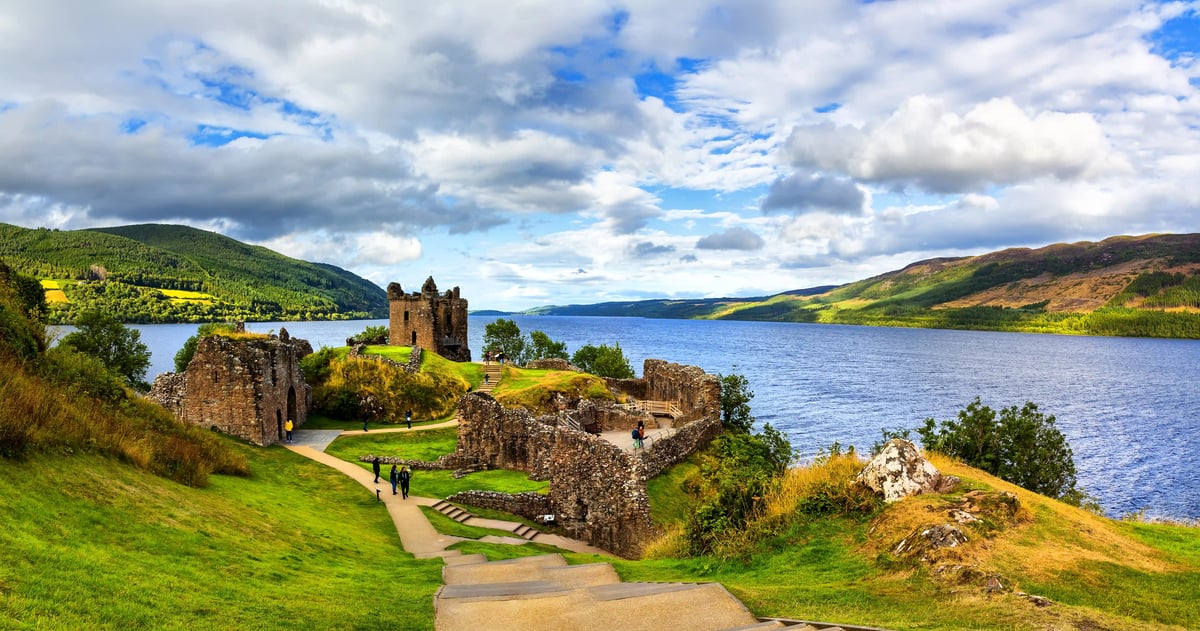 Mittelalterliche Burgruine auf einer Grünfläche am Ufer von Loch Ness mit See und Hügellandschaft unter wolkigem Himmel