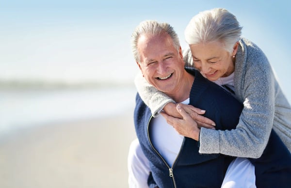 Lachendes älteres Paar am Strand, die Frau umarmt den Mann von hinten bei hellem Tageslicht und ruhiger Meereskulisse