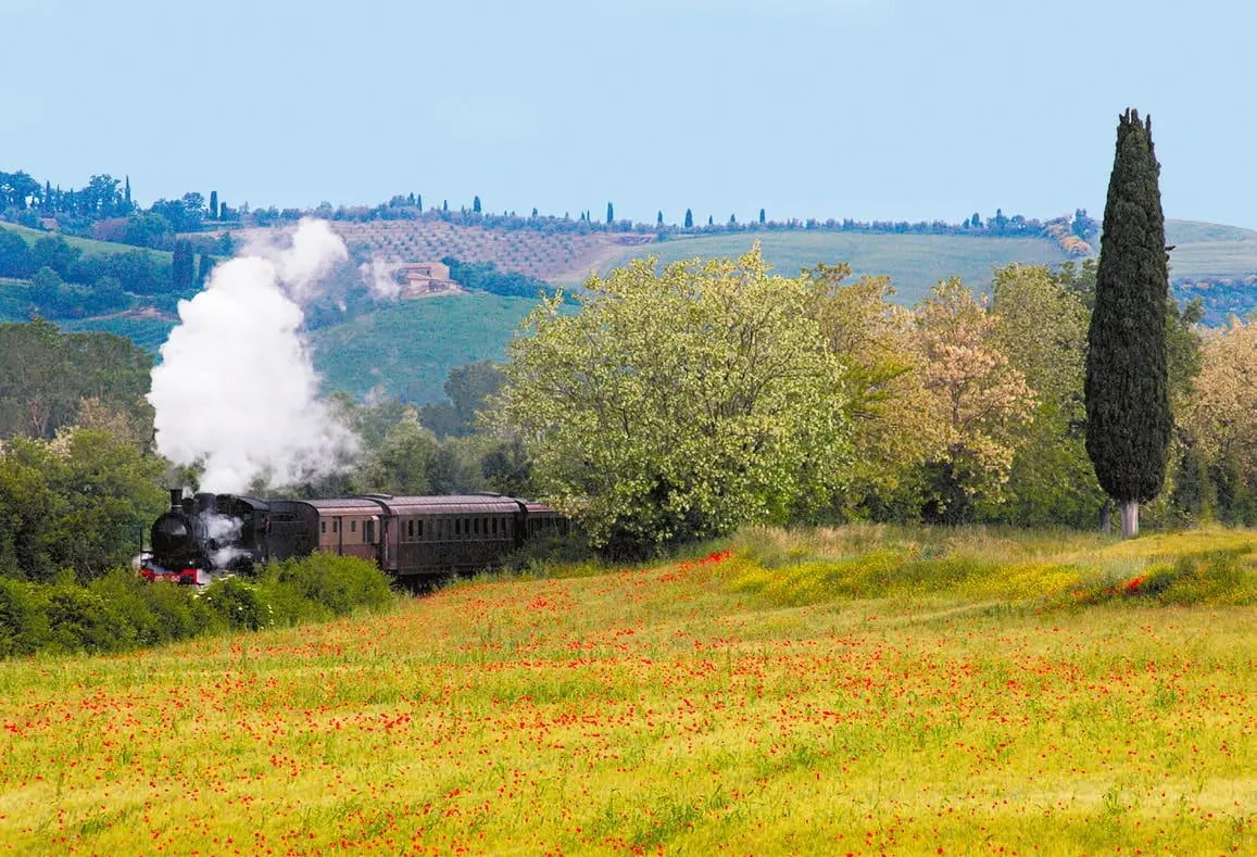 Historischer Dampfzug fährt durch blühende Landschaft der Südtoskana mit Feldern und Zypressen
