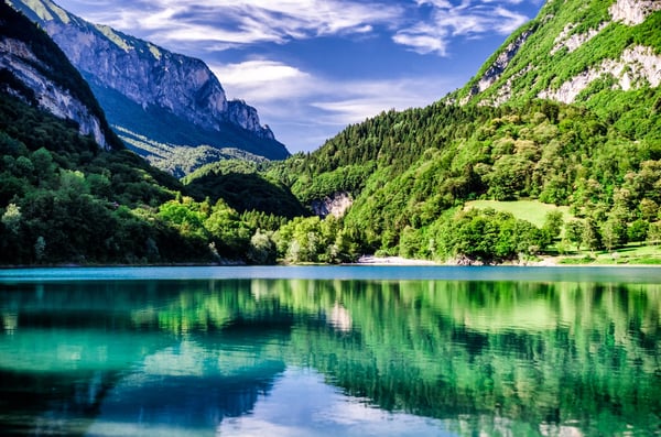 Smaragdgrüner Bergsee Lago di Tenno mit bewaldeten Hängen, klaren Spiegelungen und ruhiger Landschaft unter blauem Himmel.