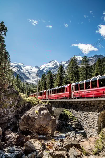 Roter Panoramazug Bernina Express fährt über eine Steinbrücke durch alpine Landschaft mit Felsen, Wäldern und schneebedeckten Gipfeln.