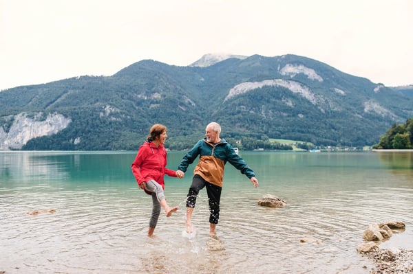 Älteres Paar steht lachend barfuss im klaren See, hält sich an den Händen und plantscht im Wasser vor grünen Bergen.