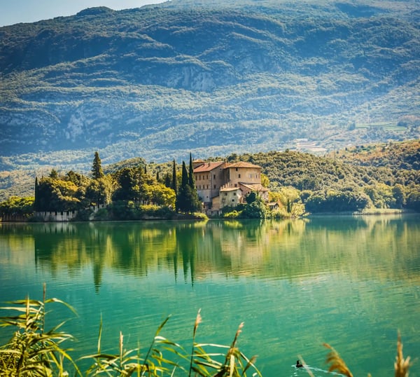 Historisches Wasserschloss liegt auf einer kleinen Insel im Lago di Toblino, umgeben von grünen Hügeln und Spiegelungen im ruhigen Wasser.