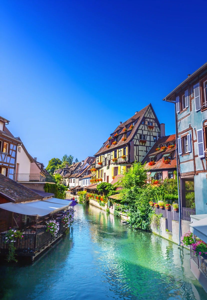Schmaler Kanal mit klarem Wasser, umgeben von Fachwerkhäusern mit Blumen und Terrassen in Colmar