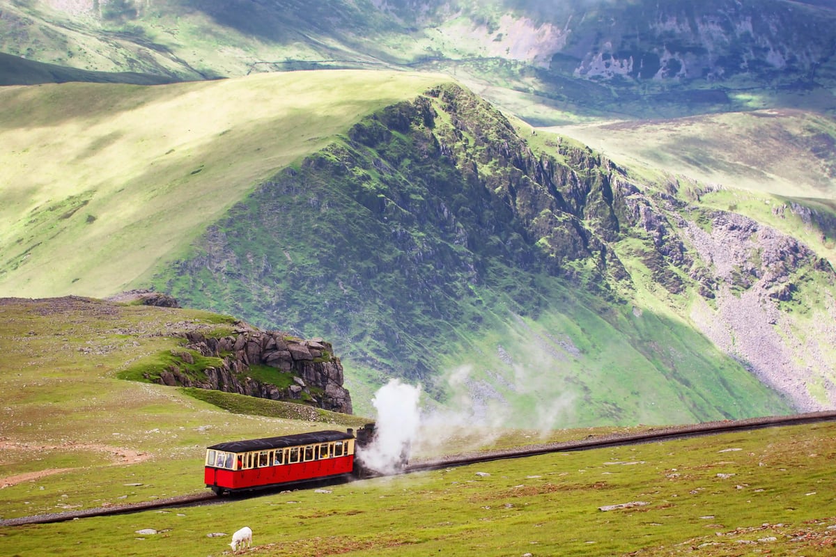 Train rouge de montagne montant a travers des collines vertes, nuage de vapeur pres des rails et pentes abruptes en arriere plan