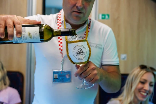 Mann schenkt Weisswein in ein Glas während einer Weinverkostung im Zug, Gäste sitzen im Hintergrund