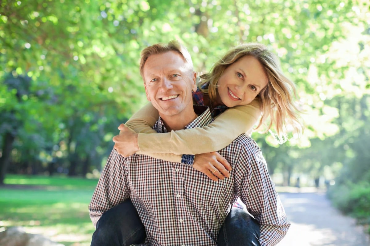 Couple souriant dans un parc verdoyant, la femme enlace lhomme par derrière, ambiance naturelle et lumière douce