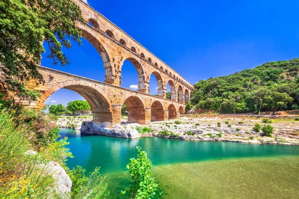 Römisches Aquädukt Pont du Gard mit mehreren Steinbögen über einem türkisfarbenen Fluss, umgeben von grüner Landschaft und blauem Himmel