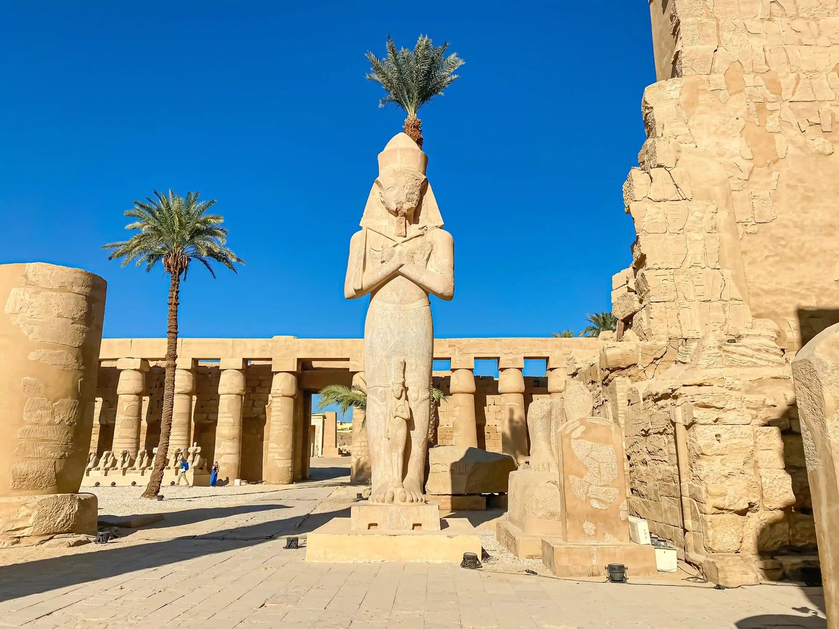 Grosse antike Statue in einer ägyptischen Tempelanlage mit Säulen, Palmen und blauem Himmel