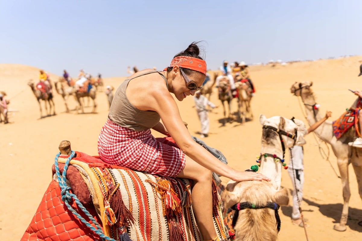 Frau reitet auf einem Kamel in der ägyptischen Wüste, umgeben von Sanddünen und weiteren Kamelen unter blauem Himmel