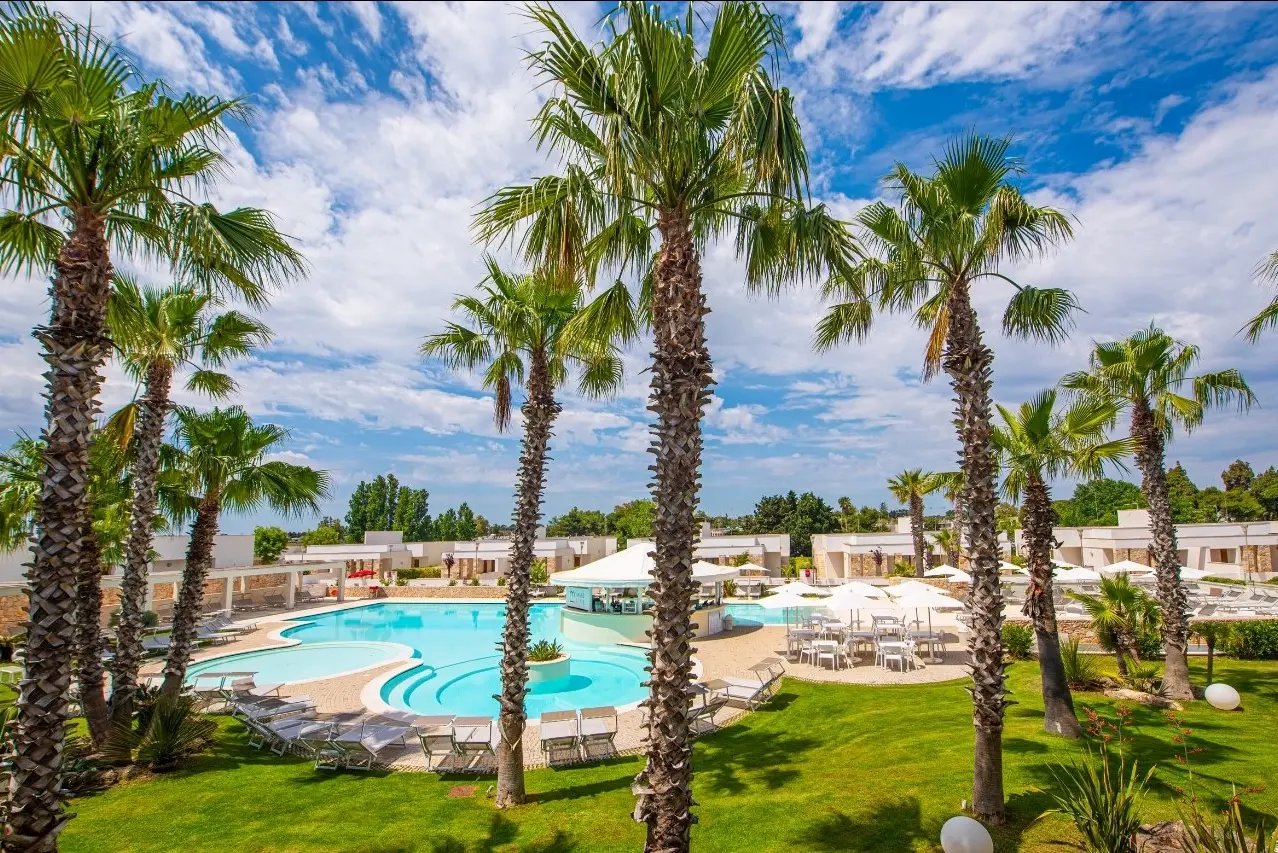 Moderne Hotelanlage mit geschwungenem Pool, Palmen, Liegestühlen und Sonnenschirmen in gepflegter Gartenanlage unter blauem Himmel