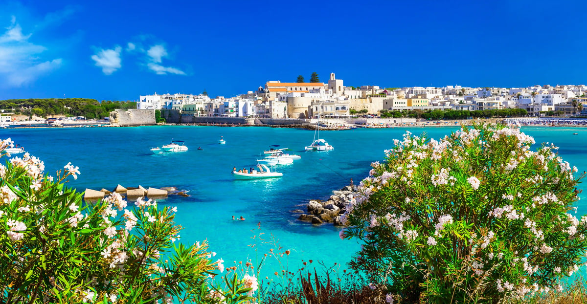 Blick auf die Küste von Otranto mit türkisfarbenem Meer, Booten, Altstadt und blühenden Sträuchern im Vordergrund