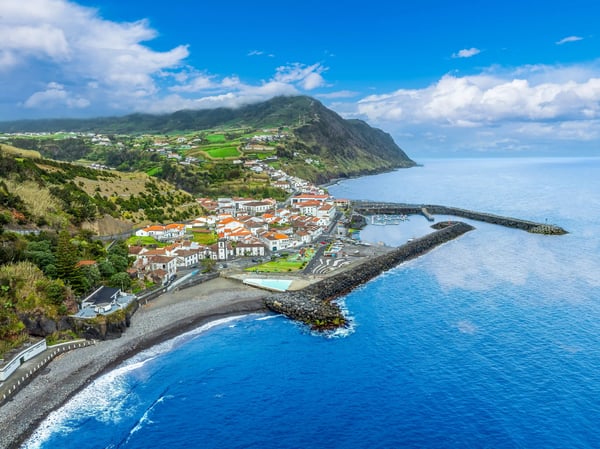 Küstendorf mit rotdachigen Häusern, Hafenanlage und blauem Atlantik vor grünen Berghängen