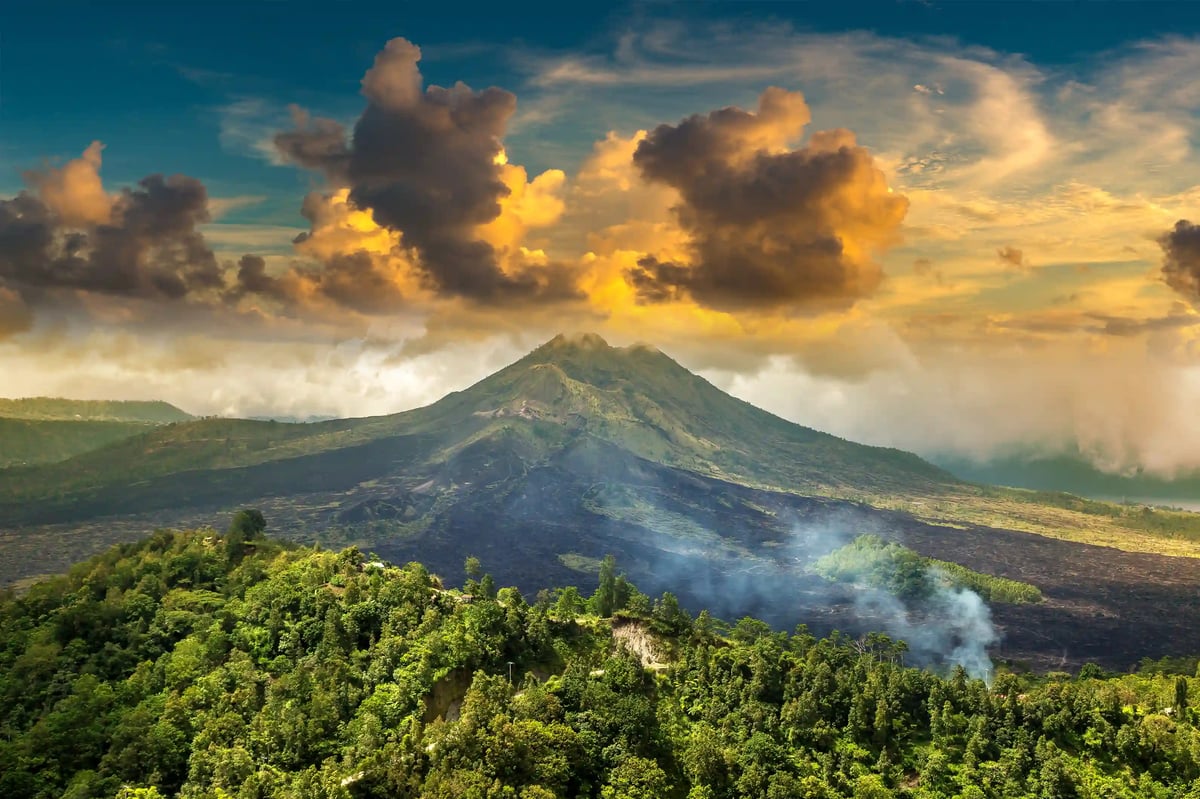 Vulkan Batur mit bewaldeter Landschaft, Rauchschwaden und dramatischem Wolkenhimmel
