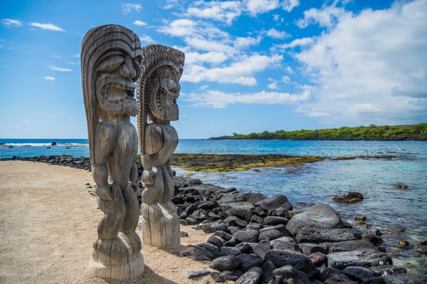 Traditionelle Holzfiguren an einer felsigen Küste mit Meer und tropischer Vegetation im Hintergrund
