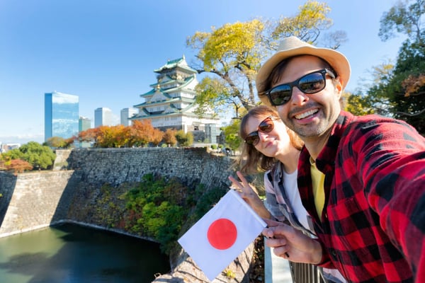 Zwei Reisende machen ein Selfie vor Osaka Castle und halten eine japanische Flagge