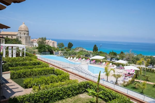 Hotelanlage in Tropea mit grossem Poolbereich, Liegestühlen und Sonnenschirmen sowie Blick auf das türkisfarbene Meer und historische Gebäude im Hintergrund