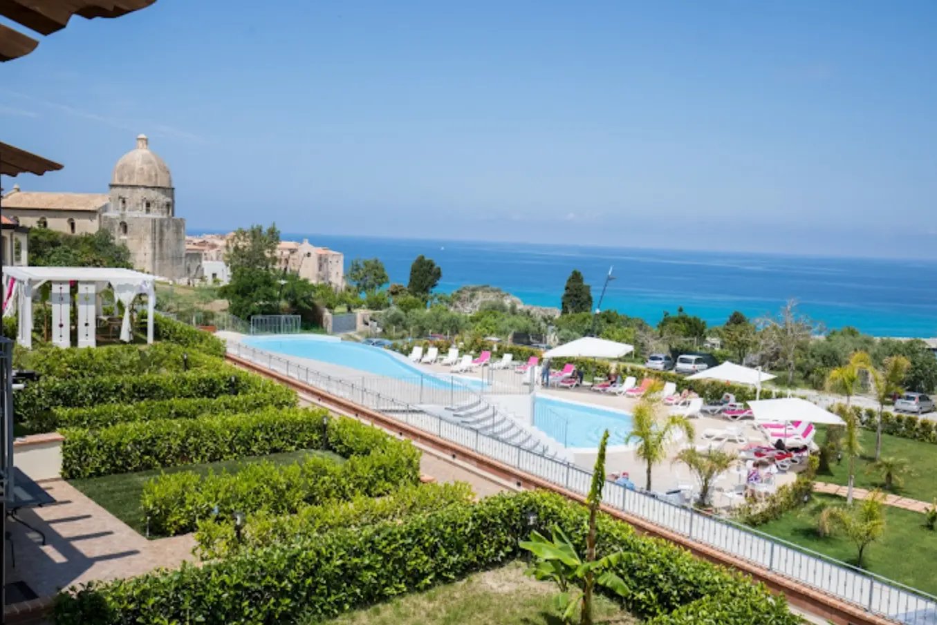 Hotelanlage in Tropea mit grossem Poolbereich, Liegestühlen und Sonnenschirmen sowie Blick auf das türkisfarbene Meer und historische Gebäude im Hintergrund