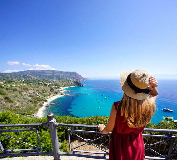 Frau mit Strohhut blickt von einer Aussichtsplattform auf türkisblaues Meer, felsige Küste und grüne Hügel in Kalabrien