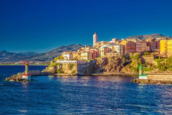 Blick auf Bastia mit farbigen Häusern auf einem Felsen, Leuchttürmen im Hafen und blauem Meer