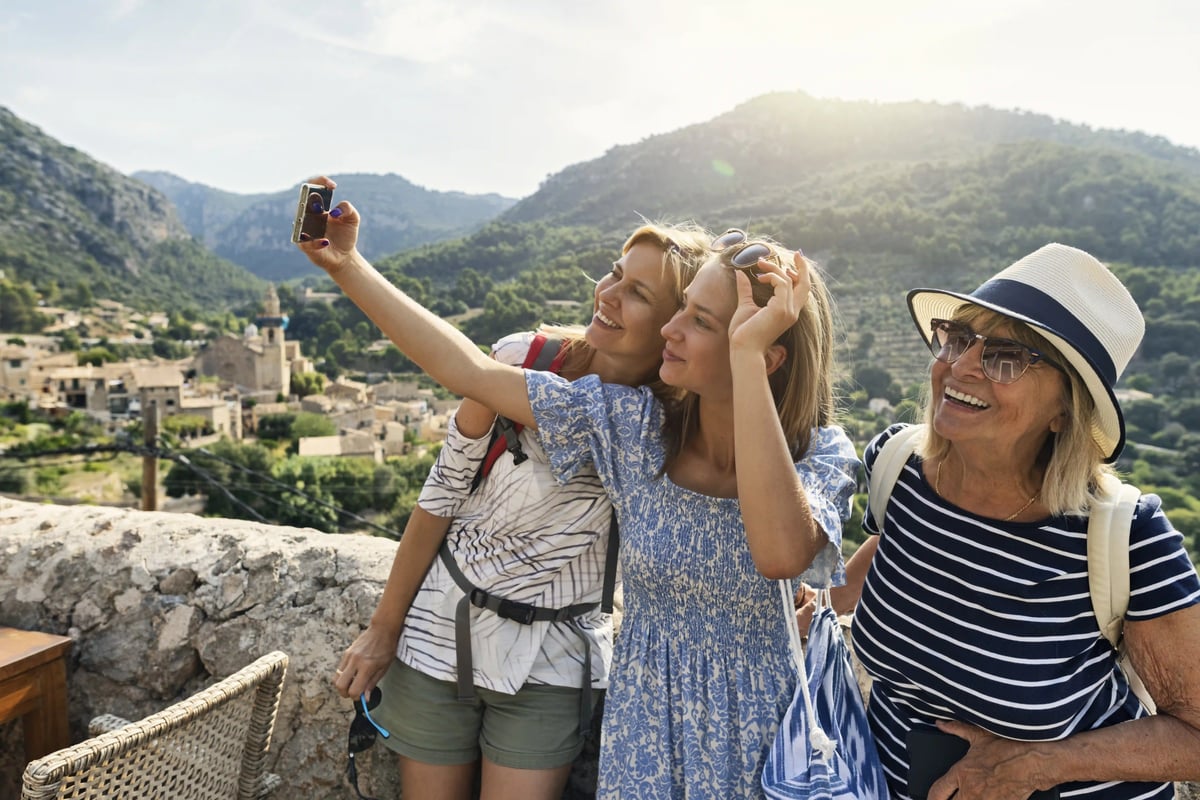 Drei Frauen machen ein Selfie an einem Aussichtspunkt mit Dorf und Bergen im Hintergrund