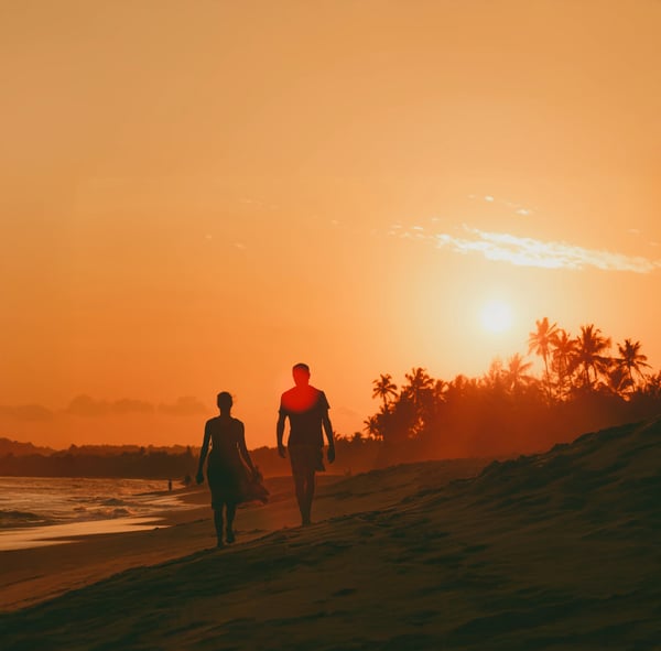 Zwei Personen spazieren bei Sonnenuntergang am Strand entlang, mit Palmen und orangefarbenem Himmel