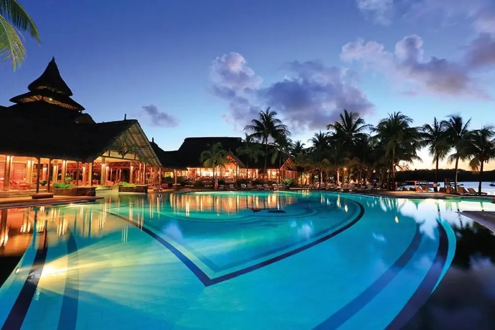 Beleuchteter Poolbereich eines Resorts bei Abenddämmerung, umgeben von Palmen und Gebäuden