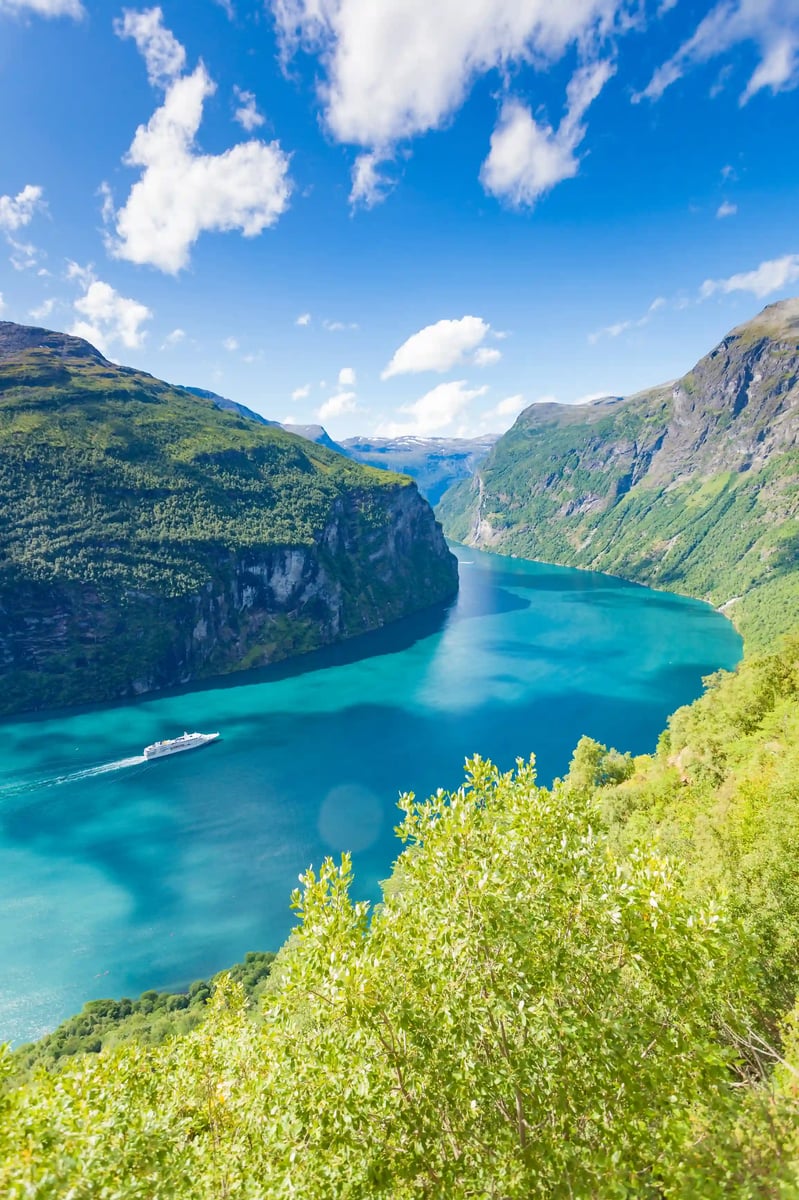 Blick auf den türkisblauen Geirangerfjord mit steilen grünen Bergen und einem Kreuzfahrtschiff auf dem Wasser