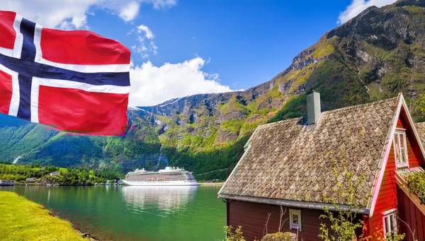 Rotes Holzhaus am Fjord mit wehender norwegischer Flagge und Kreuzfahrtschiff im Hintergrund