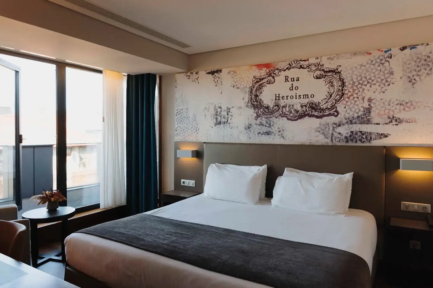 Chambre d'hôtel moderne avec un lit double, des murs décorés, des tables de chevet avec lampes et de grandes fenêtres équipées de rideaux