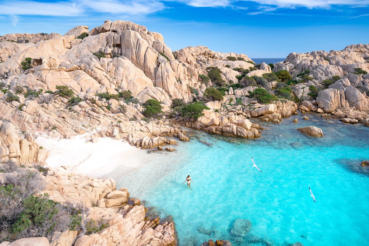 Felsige Bucht mit türkisfarbenem Meer, grüner Vegetation und Badegästen an der Küste Sardiniens