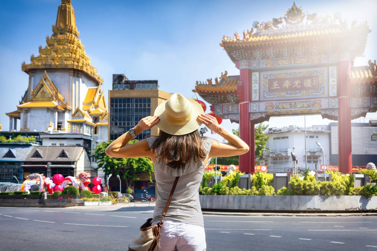 Frau mit Strohhut steht auf einer Strasse und blickt auf eine farbenreiche Tempelanlage mit chinesischem Torbogen in Bangkok