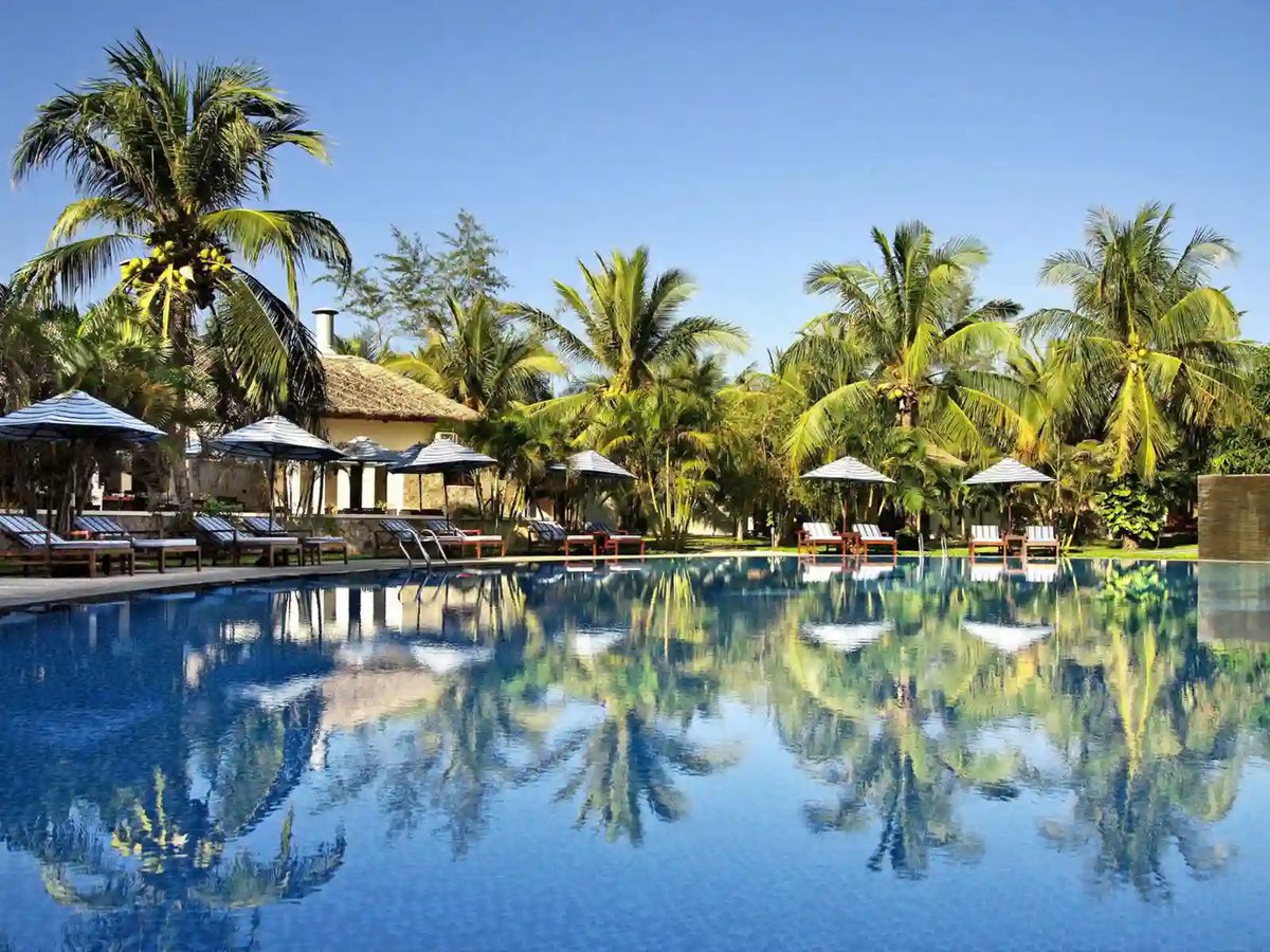 Hotelpool mit Palmen, Liegen und Spiegelung vor tropischer Küstenlandschaft in Mui Ne