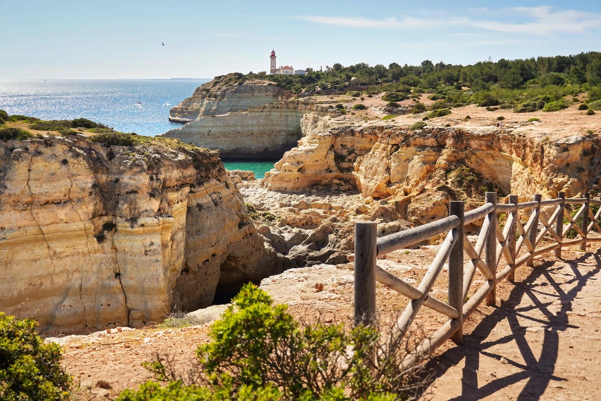 Klippenlandschaft mit Wanderweg, Holzgeländer und Leuchtturm auf einer Felsnase über dem Meer