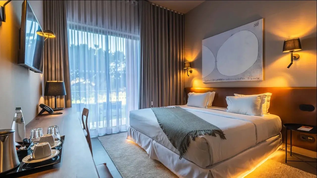 Modernes Hotelzimmer mit Doppelbett, warmer Beleuchtung, Schreibtisch, Balkonfenster mit Vorhängen und stilvoller Wanddekoration.