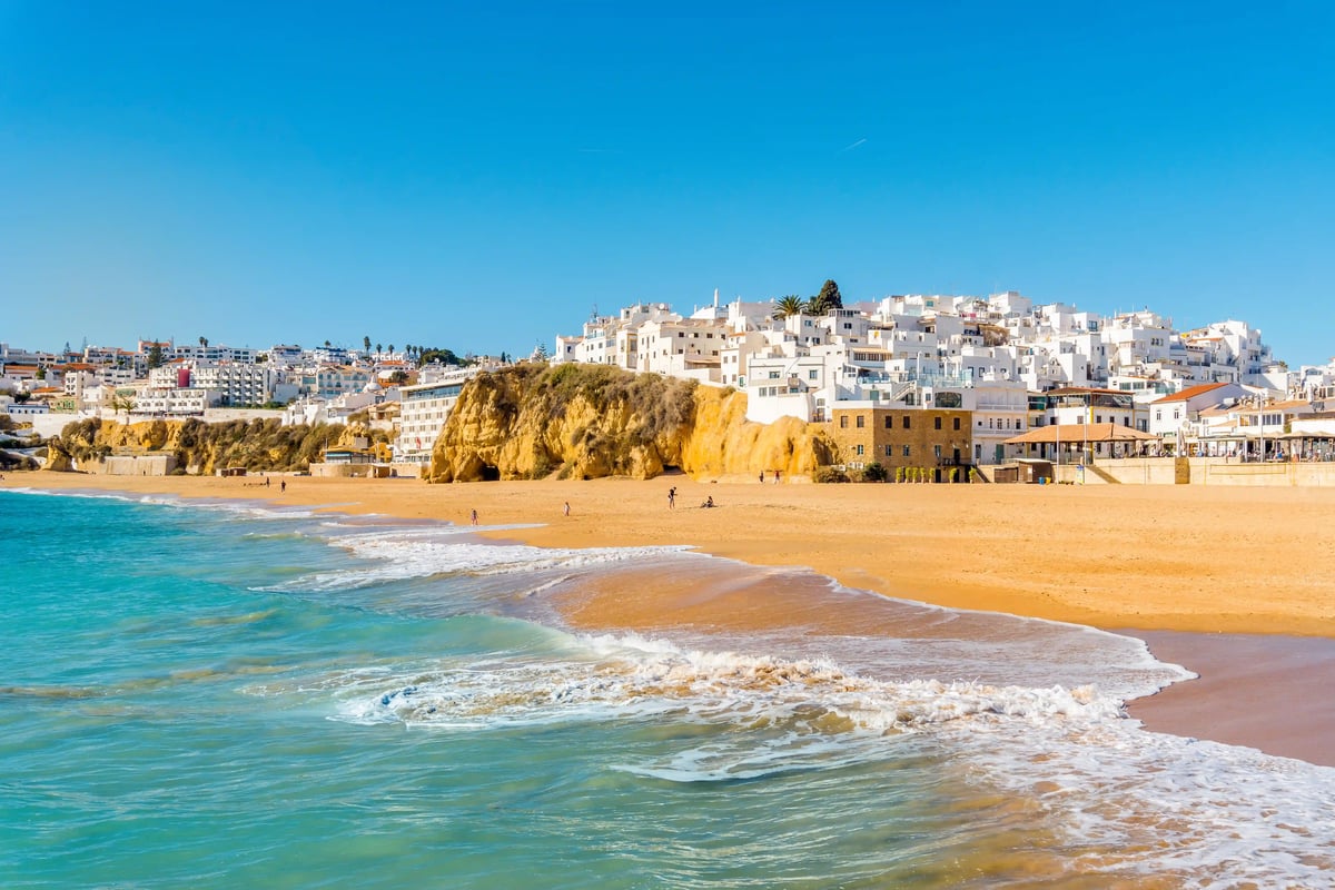 Breiter Sandstrand mit sanften Wellen, türkisem Meer und weissen Häusern auf einer Felsküste im Hintergrund
