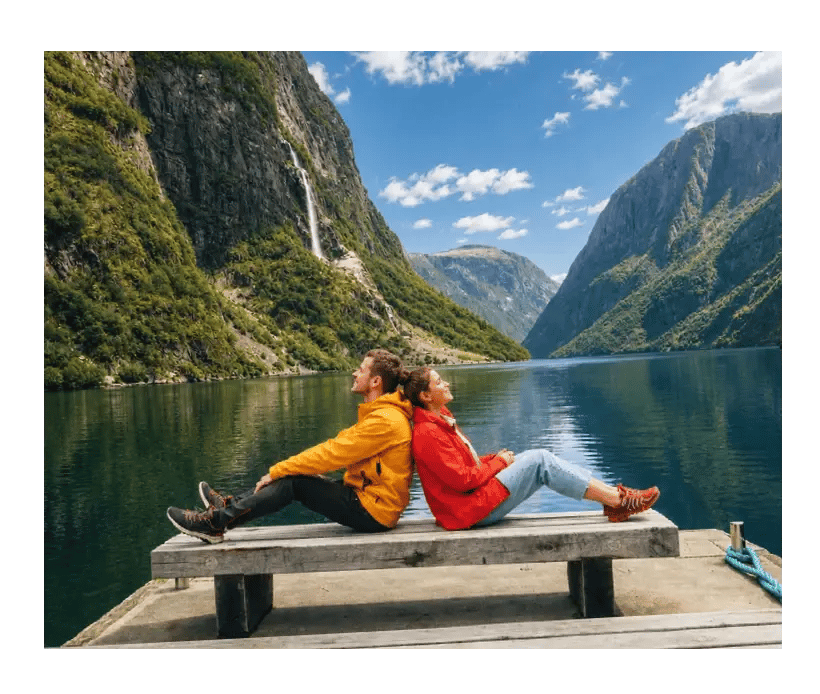Zwei Wandernde entspannen auf einem Steg vor einem norwegischen Fjord mit Wasserfall.