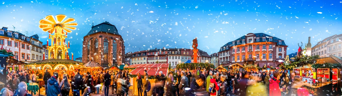 Ein belebter Weihnachtsmarkt in einer historischen Altstadt bei Dämmerung unter leichtem Schneefall, mit einer grossen Weihnachtspyramide links und festlich beleuchteten Ständen.