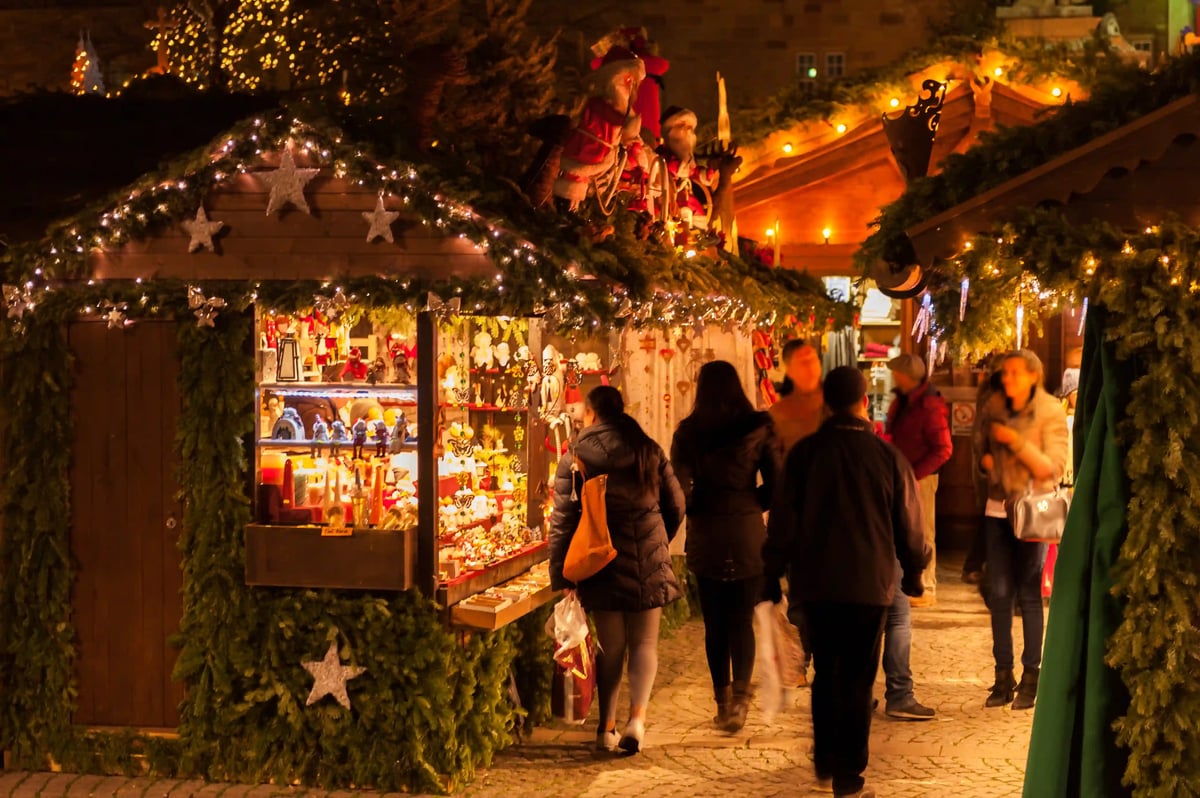 Festlich dekorierte und warm beleuchtete Holzhütten auf einem Weihnachtsmarkt bei Nacht, an denen Passanten vorbeigehen.