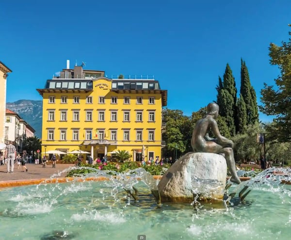 Brunnen mit Statue auf einem Platz in Riva del Garda vor einem gelben Hotelgebäude