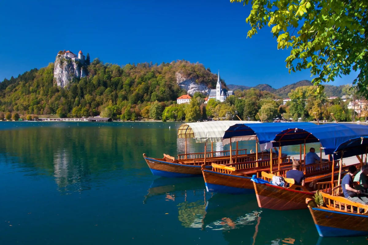 Traditionelle Holzboote am Ufer des Bleder Sees mit Kirche und Burg auf bewaldeten Huegeln im Hintergrund