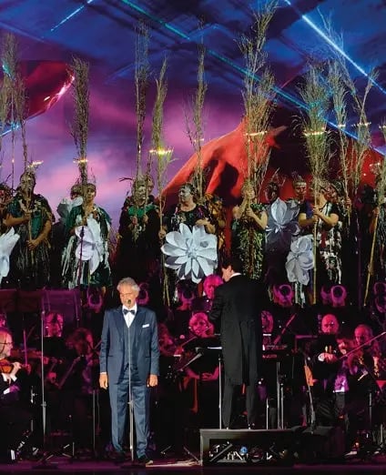 Andrea Bocelli steht auf grosser Open Air Bühne vor Orchester, Chor und farbig beleuchteter Kulisse bei Abendstimmung