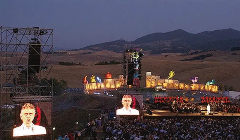 Grand théâtre en plein air dans un paysage vallonné avec scène, public et éclairages à la tombée du jour