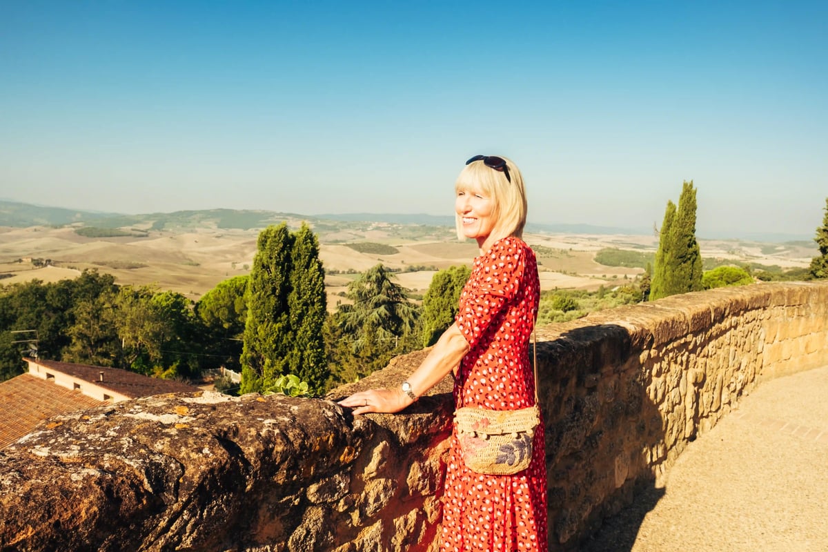 Femme souriante debout contre un mur en pierre regardant un vaste paysage vallonné de Toscane sous le soleil