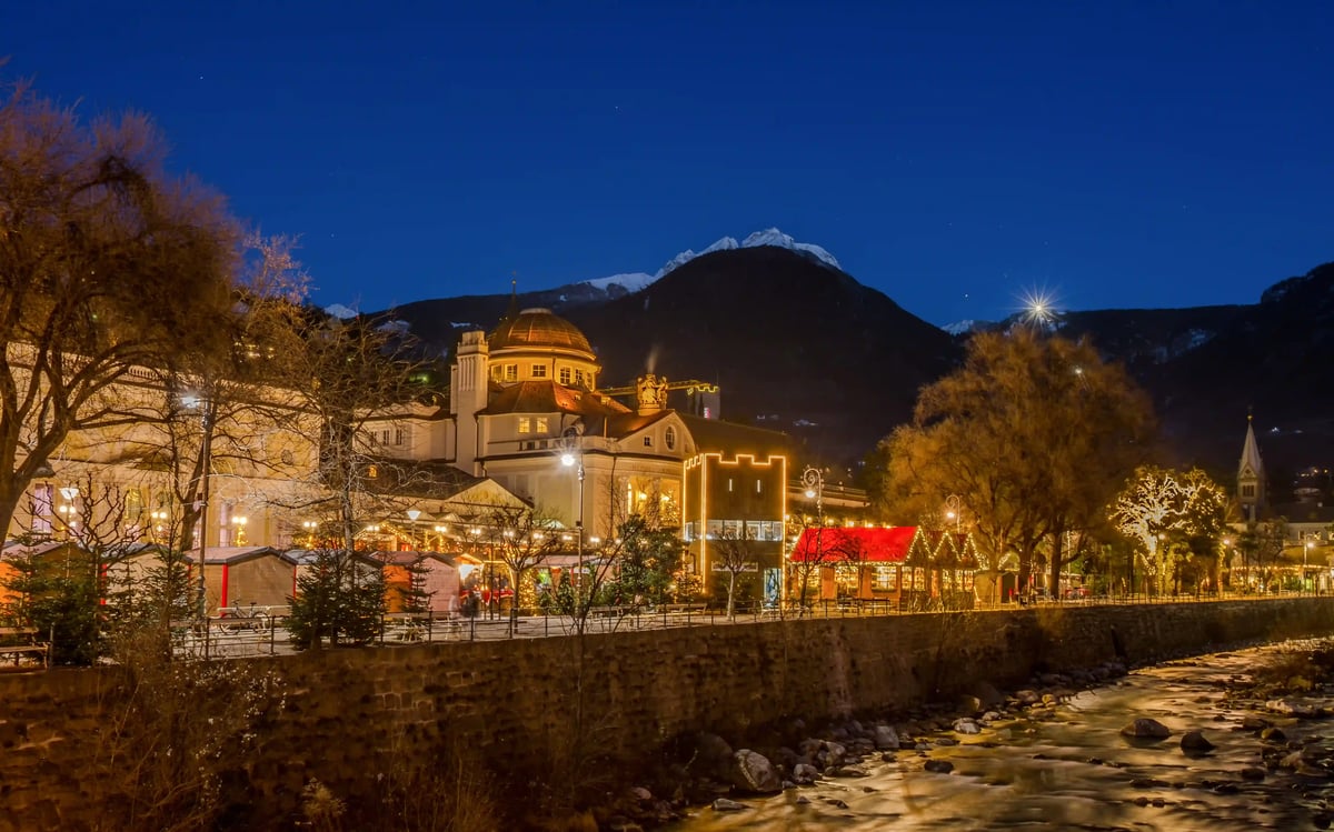 Weihnachtlich beleuchtete Promenade in Meran mit Adventsmarkt, Fluss und Bergen bei Nacht.