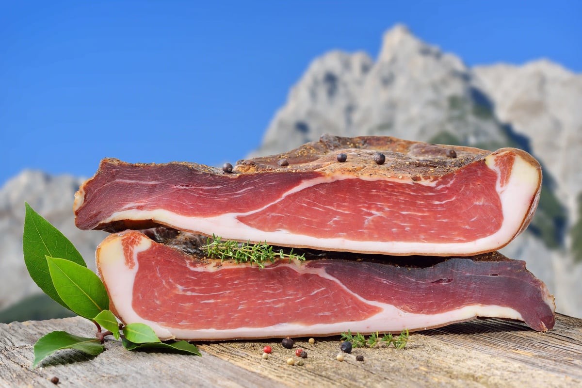Südtiroler Speck in Scheiben auf Holztisch mit Kräutern vor Bergkulisse
